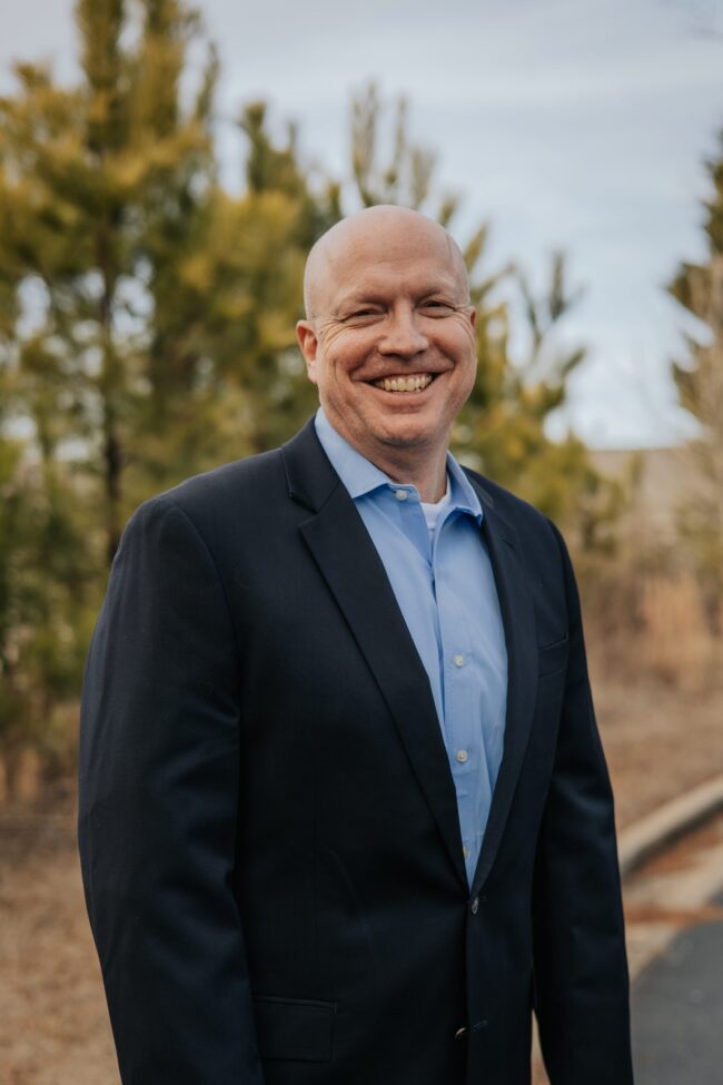 A smiling man in a dark suit standing outdoors with trees in the background.