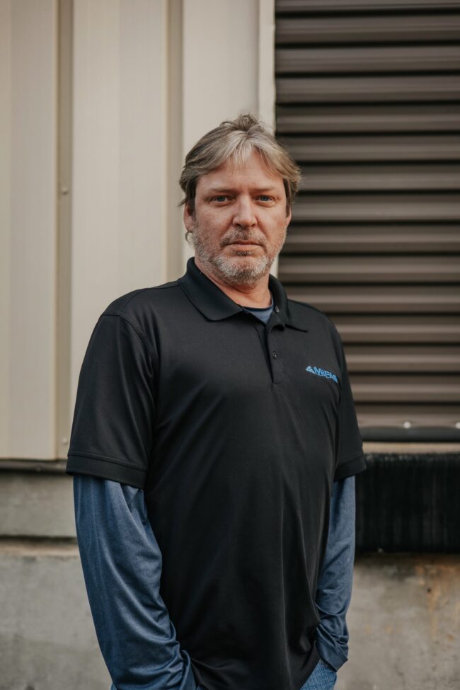 A man in a black polo shirt stands outdoors against a building wall.