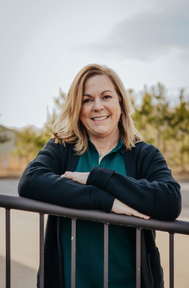 Smiling woman leaning on a railing outdoors with trees in the background.