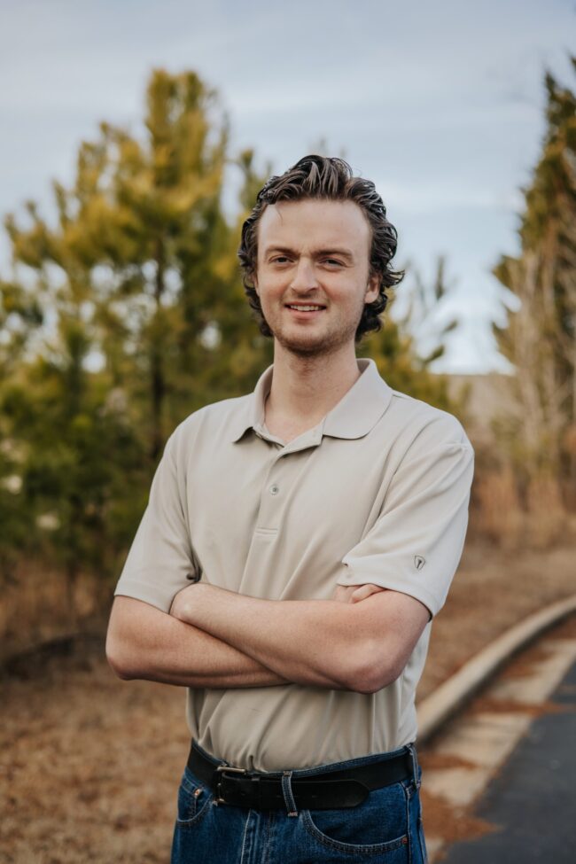 Young man smiling with arms crossed near railroad tracks.